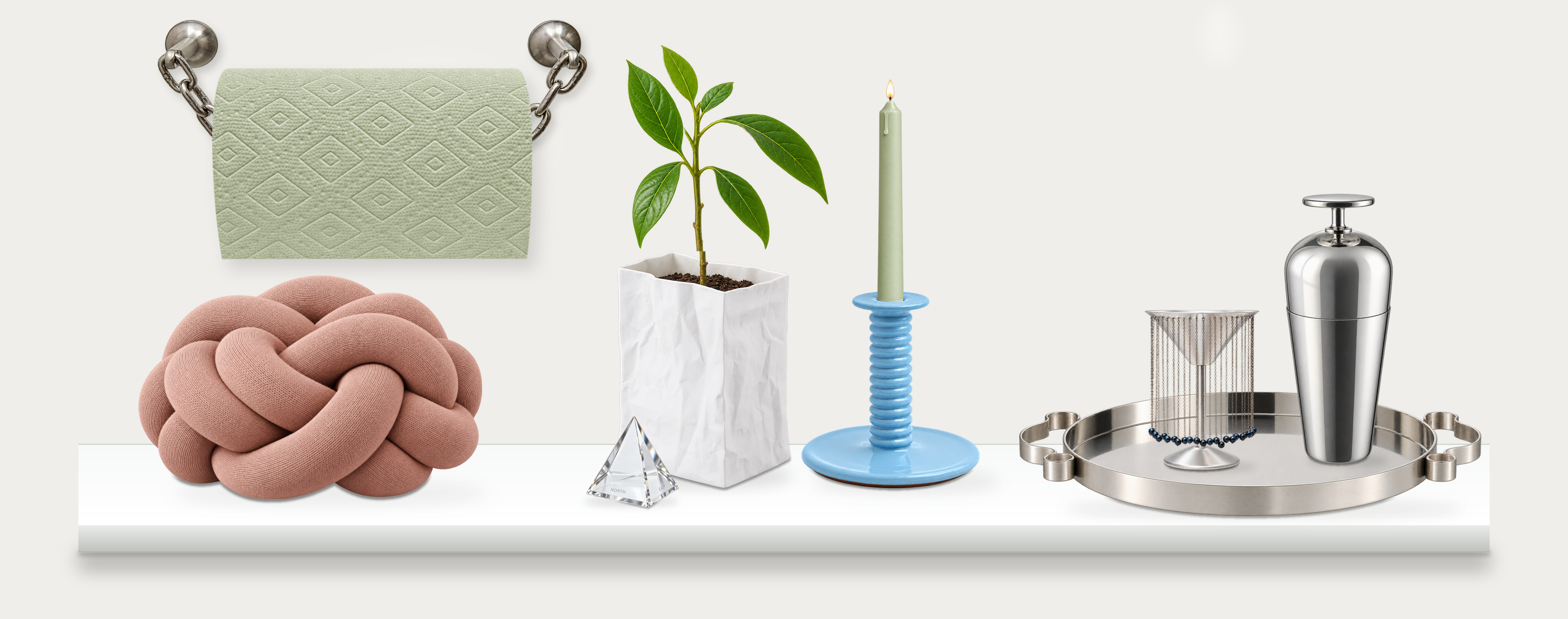 A white shelf with sage paper towels on silver chains, a dusty pink knot cushion, a crumpled white Räder porcelain bag vase with an avocado sapling and a small crystal compass pyramid in front of its left corner, a blue candleholder with a lit sage taper, and a silver tray holding a fringed cup and sculptural Tending Box shaker.